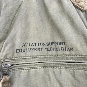 Puede incluir: Primer plano de tela verde oliva con el texto "AVIATION SUPPORT EQUIPMENT TECHNICIAN" impreso en negro. Una cremallera y tela beige son visibles en la parte inferior y superior de la imagen.