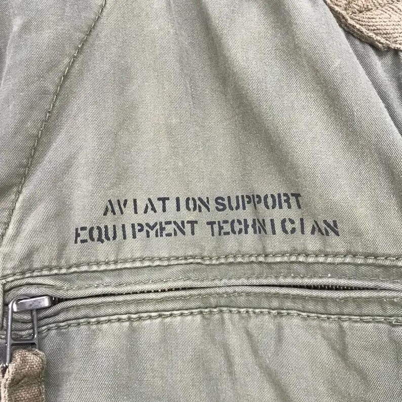 Puede incluir: Primer plano de tela verde oliva con el texto "AVIATION SUPPORT EQUIPMENT TECHNICIAN" impreso en negro. Una cremallera y tela beige son visibles en la parte inferior y superior de la imagen.