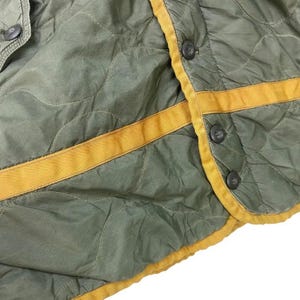 Op de afbeelding: Close-up van een gewatteerd olijfgroen kledingstuk met gele bies en donkere knopen. De stof heeft een ruitpatroon. De gele bies omzoomt de randen en loopt over de voorkant. De knopen zijn gelijkmatig verdeeld.