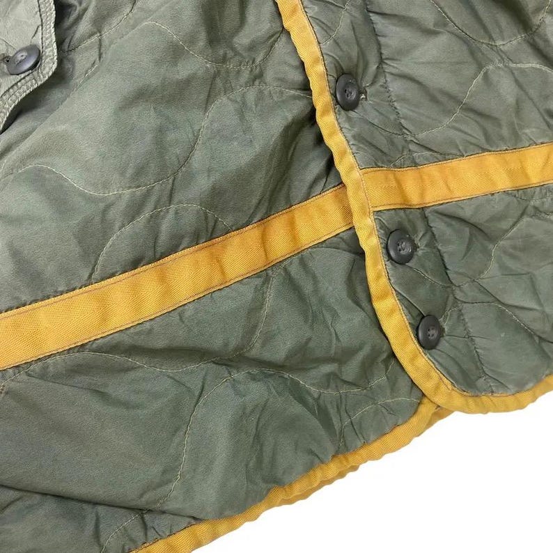 Op de afbeelding: Close-up van een gewatteerd olijfgroen kledingstuk met gele bies en donkere knopen. De stof heeft een ruitpatroon. De gele bies omzoomt de randen en loopt over de voorkant. De knopen zijn gelijkmatig verdeeld.