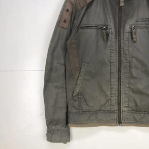Puede incluir: Chaqueta verde oliva con cremallera completa, m&uacute;ltiples bolsillos y detalles marrones en los hombros y las mangas. La chaqueta tiene un dise&ntilde;o de inspiraci&oacute;n militar con un aspecto desgastado. El texto "AVIATION EQUIPMENT" est&aacute; impreso en el pecho derecho.