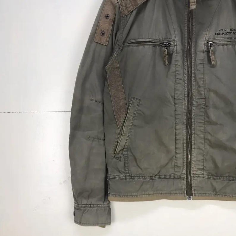 Puede incluir: Chaqueta verde oliva con cremallera completa, m&uacute;ltiples bolsillos y detalles marrones en los hombros y las mangas. La chaqueta tiene un dise&ntilde;o de inspiraci&oacute;n militar con un aspecto desgastado. El texto "AVIATION EQUIPMENT" est&aacute; impreso en el pecho derecho.