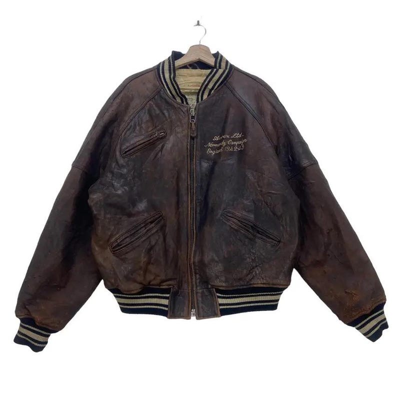 May include: A vintage brown leather bomber jacket with a zip closure, two front pockets, and ribbed cuffs and hem. The jacket has the embroidered text "Storm Ltd. Normandy Company England 1944-1945" on the chest.
