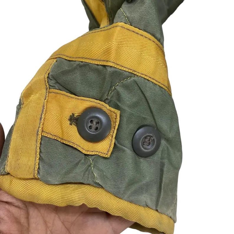 Op de afbeelding: Close-up van een deel van een gewatteerde jas. De jas heeft olijfgroene stof met gele randen en accenten. Twee donkergrijze knopen zijn zichtbaar, &eacute;&eacute;n op een gele vierkante patch. De stiksels zijn zichtbaar.