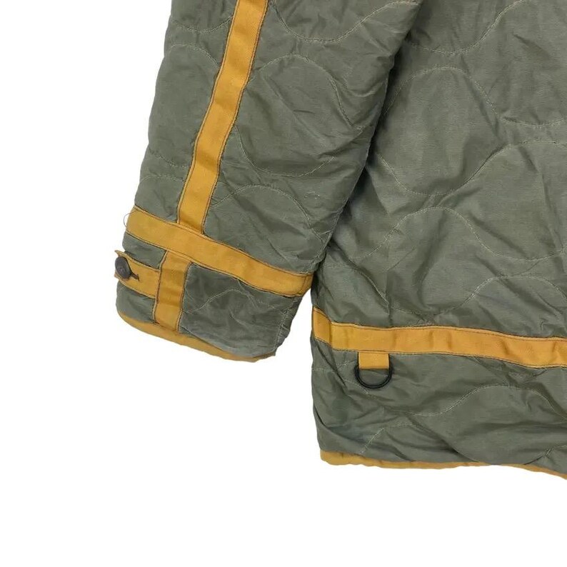 Op de afbeelding: Close-up van een gewatteerde olijfgroene jas met gele strepen en bies. De jas heeft een ruitpatroon en een knoopsluiting op de mouw. Een metalen D-ring is zichtbaar aan de onderrand.