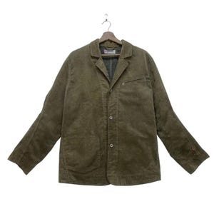 Puede incluir: Chaqueta de pana verde oliva con solapas con muescas, tres botones y dos bolsillos delanteros. La chaqueta está colgada de una percha de madera sobre un fondo blanco. Las mangas son largas y la chaqueta parece de peso medio.