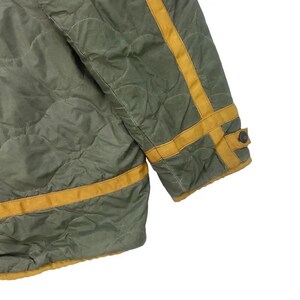 Op de afbeelding: Close-up van een gewatteerde olijfgroene jas met gele bies. De jas heeft een ruitpatroon en een manchet met knoop. De gele bies zit op de mouwen en langs de randen van de jas.