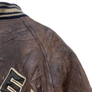 May include: Close-up of a brown leather jacket with a black and beige striped collar. The jacket shows signs of wear and tear, with a distressed look. The letter "E" is visible on the jacket.