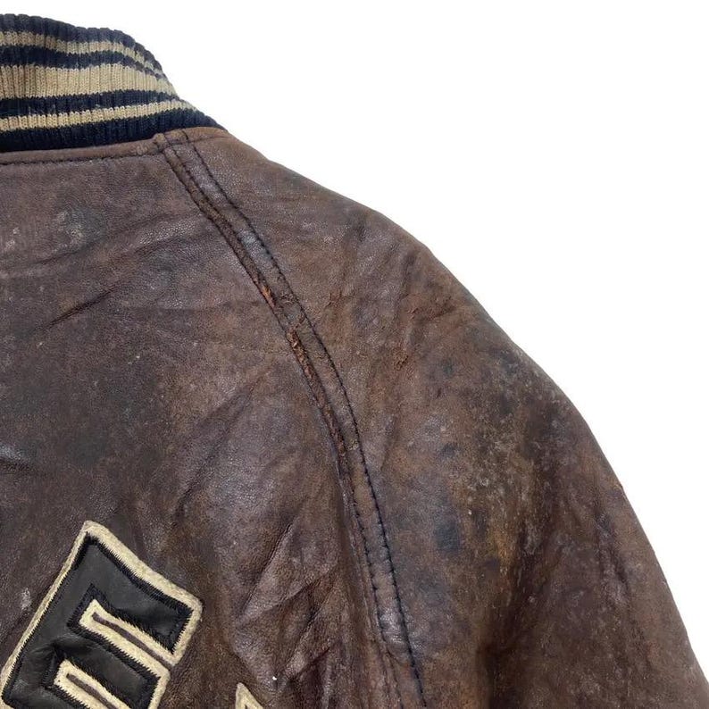 May include: Close-up of a brown leather jacket with a black and beige striped collar. The jacket shows signs of wear and tear, with a distressed look. The letter "E" is visible on the jacket.