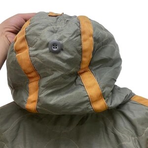 Op de afbeelding: Close-up van een gewatteerde olijfgroene capuchon met oranje bies en een grijze knoop. De capuchon maakt deel uit van een jas of jas, en de stof heeft een subtiele glans. Het ontwerp heeft een gewatteerd patroon.