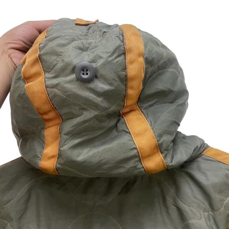 Op de afbeelding: Close-up van een gewatteerde olijfgroene capuchon met oranje bies en een grijze knoop. De capuchon maakt deel uit van een jas of jas, en de stof heeft een subtiele glans. Het ontwerp heeft een gewatteerd patroon.