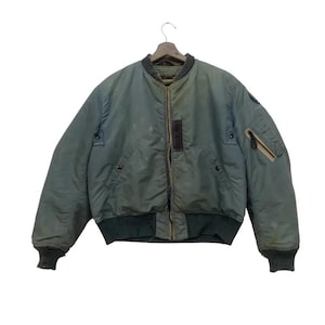 May include: An olive green bomber jacket with a brown leather patch and a zipped pocket on the left sleeve. The jacket has a ribbed collar, cuffs, and hem. It is suspended from a wooden hanger.