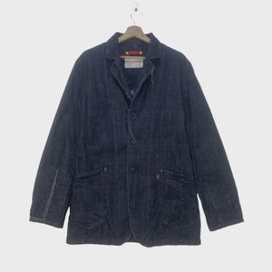 May include: Dark blue denim jacket with a classic collar, three buttons, and two front pockets. White stitching accents the seams. A red tag is visible on the pocket. The jacket is hanging on a wooden hanger.
