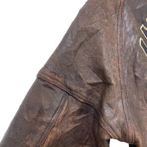 May include: Close-up of a distressed brown leather jacket. The jacket shows signs of wear and tear, with visible stitching and a worn texture. The image highlights the jacket's vintage aesthetic and rugged appeal.