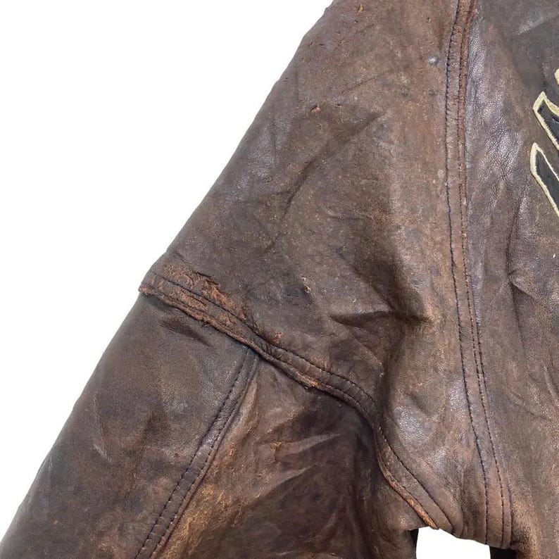 May include: Close-up of a distressed brown leather jacket. The jacket shows signs of wear and tear, with visible stitching and a worn texture. The image highlights the jacket's vintage aesthetic and rugged appeal.