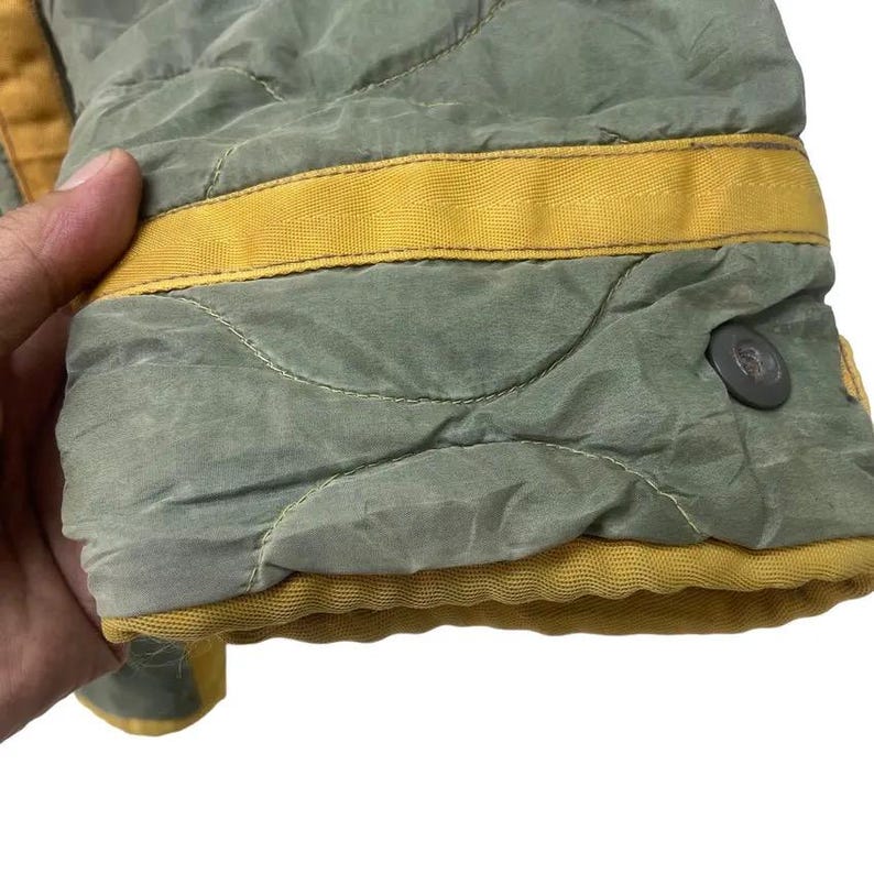 Op de afbeelding: Detail van een gewatteerde jas-mouw. De jas is olijfgroen met gele biezen en een knoopsluiting. De stof heeft een licht glanzende uitstraling.