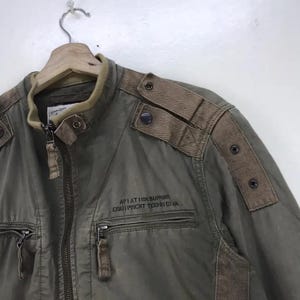 Puede incluir: Chaqueta verde oliva con cuello alzado, cierre de cremallera y m&uacute;ltiples bolsillos. Cuenta con detalles marrones en los hombros y el texto "AVIATION SUPPORT EQUIPMENT TECHNICIAN". La chaqueta est&aacute; colgada en una percha de madera.