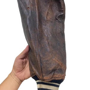 May include: Close-up of a brown leather jacket sleeve. The leather shows wear and tear, with visible stitching along the seams. The cuff is ribbed, featuring a striped pattern in navy, beige, and white.