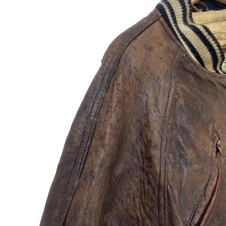 May include: Close-up of a brown leather jacket with a distressed finish. The jacket features a ribbed collar with black and beige stripes. Visible stitching details and a zipped pocket add to the vintage aesthetic. The leather has a worn, textured appearance.