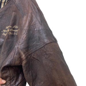May include: Close-up of a vintage brown leather jacket. The jacket has a worn, distressed appearance with visible creases and variations in colour. Gold embroidered text on the jacket reads "Apex Ltd. Normby Camp Eng 1942-43".