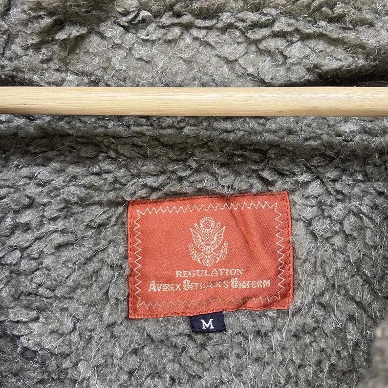 Op de afbeelding: Close-up van een grijze, getextureerde jasvoering met een rood rechthoekig label. Het label bevat de tekst "REGULATION" en "Avirex Outfitters Uniform" samen met een wapen. De jas hangt aan een houten hanger.