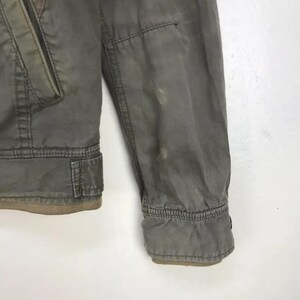 Puede incluir: Primer plano de una manga de chaqueta verde oliva descolorida. La chaqueta tiene un pu&ntilde;o acanalado y un peque&ntilde;o bolsillo en la parte superior del brazo. La tela muestra signos de desgaste, con algo de decoloraci&oacute;n.