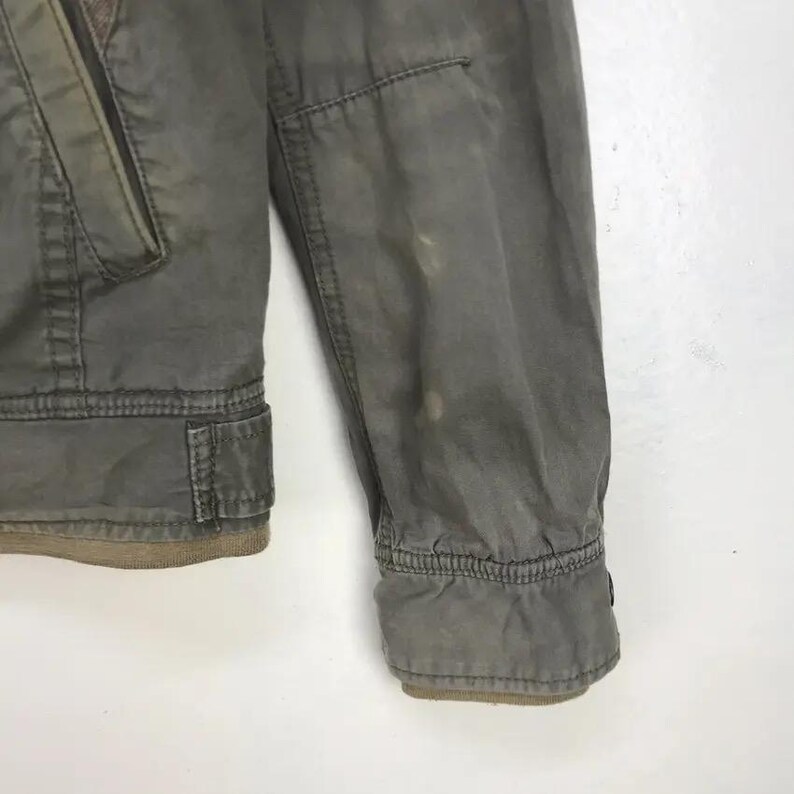 Puede incluir: Primer plano de una manga de chaqueta verde oliva descolorida. La chaqueta tiene un pu&ntilde;o acanalado y un peque&ntilde;o bolsillo en la parte superior del brazo. La tela muestra signos de desgaste, con algo de decoloraci&oacute;n.