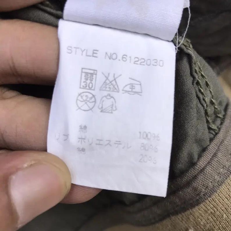 Puede incluir: Una etiqueta de ropa blanca con el texto "STYLE NO.6122030" y varios s&iacute;mbolos de cuidado. La etiqueta tambi&eacute;n enumera la composici&oacute;n de la tela: 100% algod&oacute;n, 80% poli&eacute;ster y 20% otros.