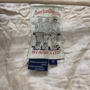 May include: A close-up of a cream-coloured quilted fabric with a rectangular tag. The tag reads "American Classics GARMENTS FOR ALL SPORTS BY AVIREX LTD." The tag also lists the materials: 100% genuine leather outshell, 50% nylon lining, and 50% acetate.