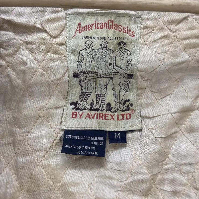 May include: A close-up of a cream-coloured quilted fabric with a rectangular tag. The tag reads "American Classics GARMENTS FOR ALL SPORTS BY AVIREX LTD." The tag also lists the materials: 100% genuine leather outshell, 50% nylon lining, and 50% acetate.