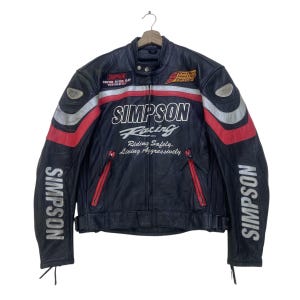 Może przedstawiać: Czarna skórzana kurtka motocyklowa z czerwonymi i srebrnymi akcentami. Kurtka posiada logo "SIMPSON Racing" oraz napis "SIMPSON" na rękawach. Widoczny jest również tekst "Riding Safely, Living Aggressively".