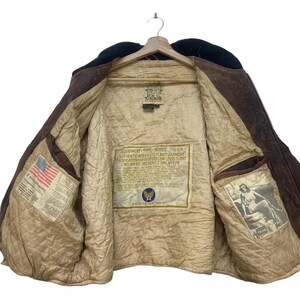 May include: A brown leather flight jacket with a black collar, displayed on a wooden hanger. The interior has a quilted lining and patches with text and illustrations. The jacket features a zip closure and several pockets.