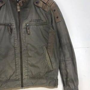 Puede incluir: Chaqueta verde oliva con cremallera completa, dos bolsillos delanteros y cuello alzado. La chaqueta tiene parches marrones en los hombros y el texto "AVIATION SUPPORT EQUIPMENT TECHNICIAN" impreso en el pecho.