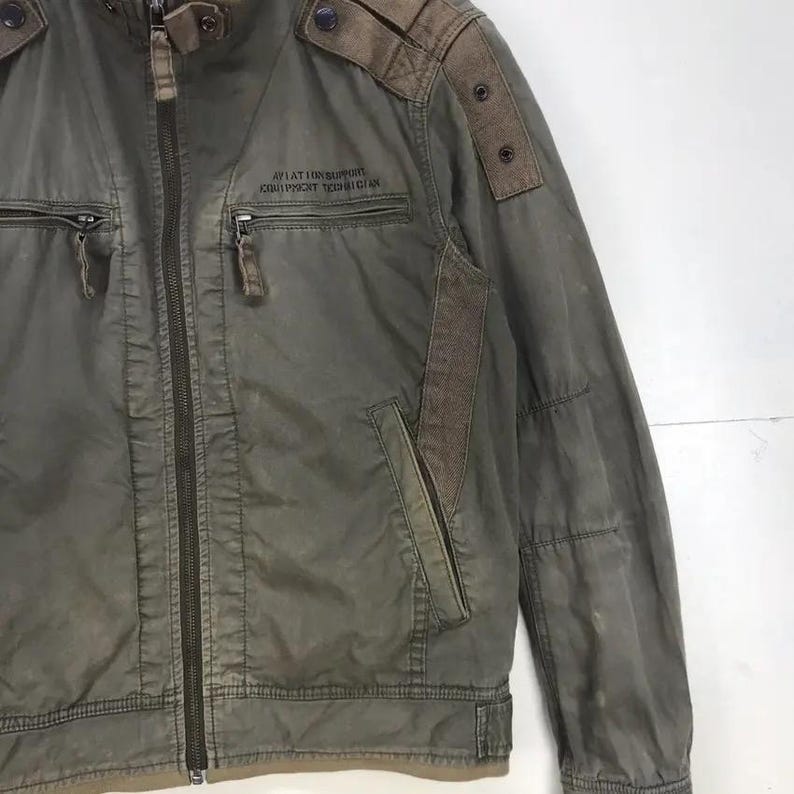 Puede incluir: Chaqueta verde oliva con cremallera completa, dos bolsillos delanteros y cuello alzado. La chaqueta tiene parches marrones en los hombros y el texto "AVIATION SUPPORT EQUIPMENT TECHNICIAN" impreso en el pecho.
