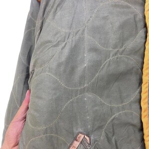 Op de afbeelding: Close-up van een gewatteerde olijfgroene jas. De jas heeft een golvend stikselpatroon en een gele bies. Een hand is zichtbaar aan de linkerkant van de afbeelding.