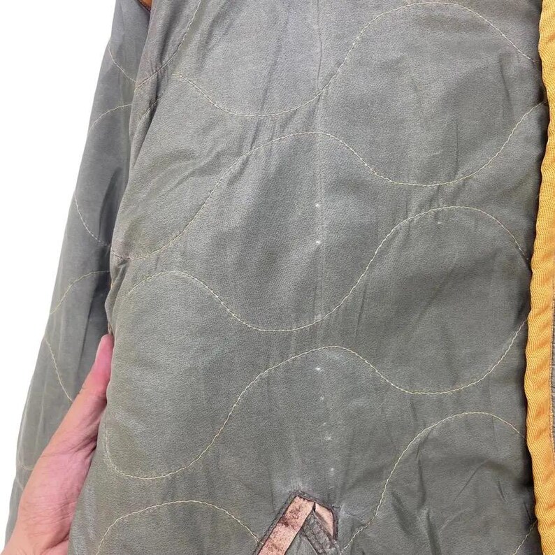 Op de afbeelding: Close-up van een gewatteerde olijfgroene jas. De jas heeft een golvend stikselpatroon en een gele bies. Een hand is zichtbaar aan de linkerkant van de afbeelding.