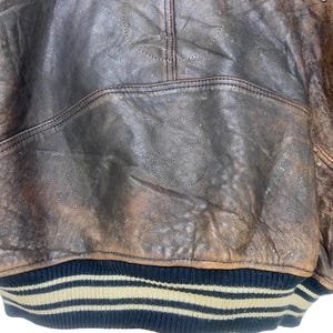 May include: Close-up of a vintage brown leather jacket. The jacket shows signs of wear and tear, with the letters "US" stitched into the leather. The bottom hem has a black and beige striped design.