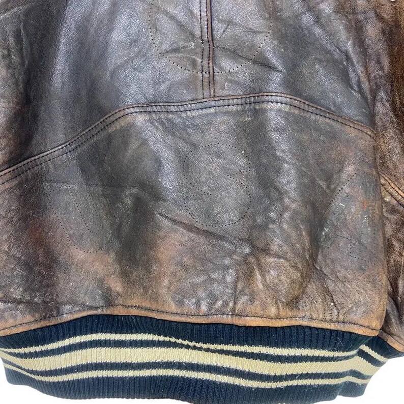 May include: Close-up of a vintage brown leather jacket. The jacket shows signs of wear and tear, with the letters "US" stitched into the leather. The bottom hem has a black and beige striped design.