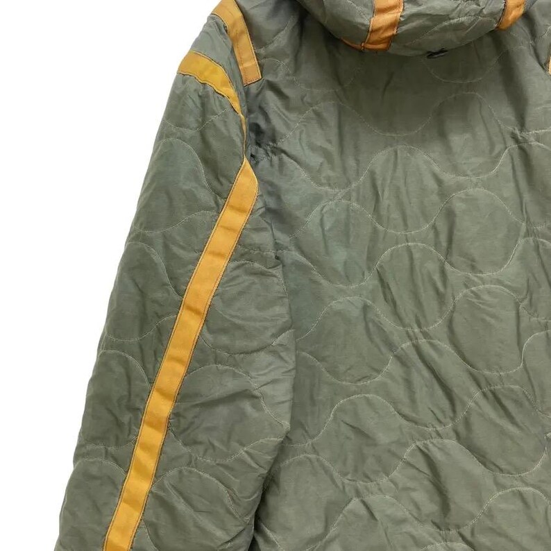 Op de afbeelding: Olijfgroene gewatteerde jas met een capuchon en gele strepen. De jas heeft een gewatteerd patroon en gele accenten langs de naden en capuchon. De jas is gemaakt van een lichtgewicht materiaal.