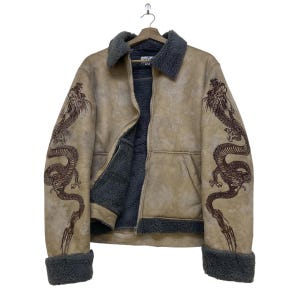 Könnte beinhalten: Eine beigefarbene Jacke mit dunkelgrauem Kunstfellfutter, Kragen und Ärmelbündchen. Die Ärmel sind mit dunkelbraunen Drachenstickereien versehen. Die Jacke verfügt über einen Frontreißverschluss und zwei seitliche Eingrifftaschen.