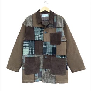 Könnte beinhalten: Eine langärmelige Jacke mit einem Patchwork-Design in verschiedenen Braun-, Beige- und Schwarztönen. Das Kleidungsstück verfügt über einen Kragen, eine Knopfleiste vorne, eine Brusttasche und zwei große untere Taschen aus verschiedenen Stoffen.