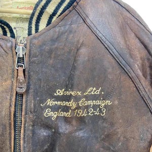 May include: Close-up of a vintage brown leather bomber jacket. The jacket has a black and tan striped collar and a zipper. Gold embroidery reads "Avirex Ltd. Normandy Campaign England 1942-43". The leather shows signs of wear and tear.