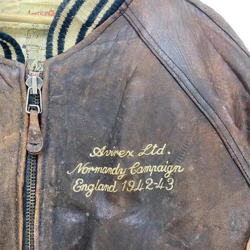 May include: Close-up of a vintage brown leather bomber jacket. The jacket has a black and tan striped collar and a zipper. Gold embroidery reads "Avirex Ltd. Normandy Campaign England 1942-43". The leather shows signs of wear and tear.
