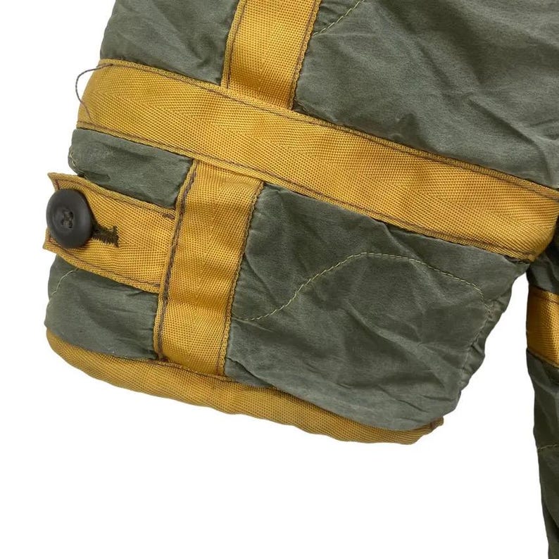 Op de afbeelding: Detail van olijfgroene stof met mosterdgele accenten. De stof is gewatteerd en heeft een knoopsluiting. Het ontwerp omvat horizontale en verticale gele banden, waardoor een rasterpatroon ontstaat.