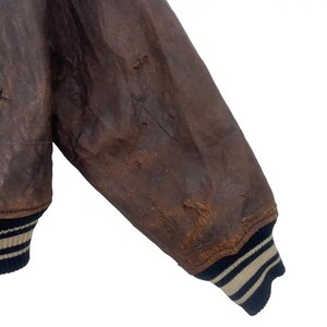 May include: Close-up of a distressed brown leather jacket sleeve. The sleeve shows wear and tear, with visible imperfections. The cuff features black and tan striped ribbing. The jacket appears vintage or pre-owned.