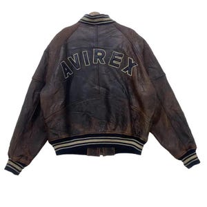 May include: Brown leather bomber jacket with the word "AVIREX" arched across the back in a cream-coloured font. The jacket has a ribbed collar, cuffs, and hem with navy blue and cream stripes. The jacket is a vintage style.
