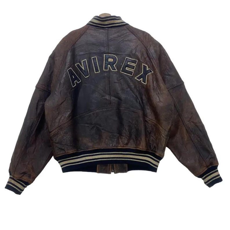 May include: Brown leather bomber jacket with the word "AVIREX" arched across the back in a cream-coloured font. The jacket has a ribbed collar, cuffs, and hem with navy blue and cream stripes. The jacket is a vintage style.