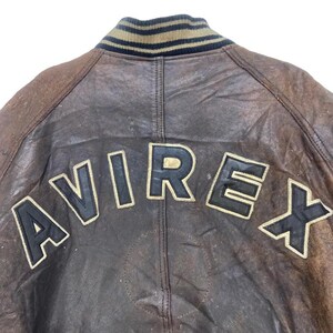 May include: Close-up of a brown leather jacket back with the brand name "AVIREX" in large, black, block letters outlined in gold. The jacket has a ribbed collar with stripes of tan, black, and navy blue.