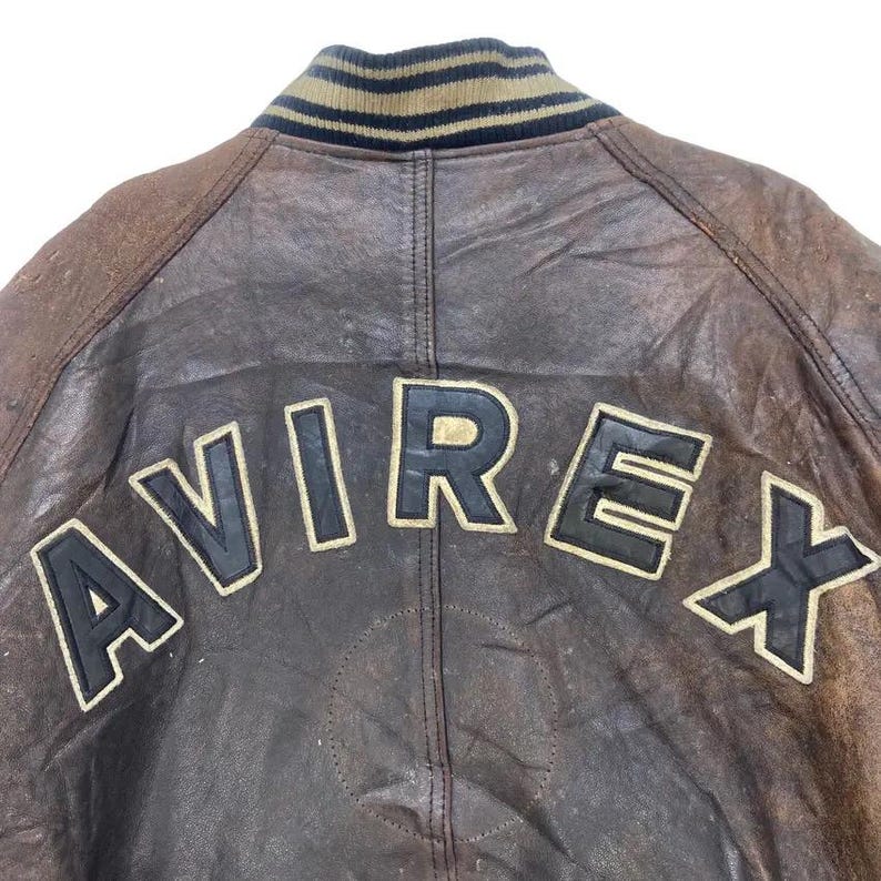 May include: Close-up of a brown leather jacket back with the brand name "AVIREX" in large, black, block letters outlined in gold. The jacket has a ribbed collar with stripes of tan, black, and navy blue.