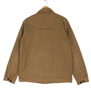 Puede incluir: Chaqueta de trabajo color marrón con el texto "ALOHA FROM HELL" impreso en la espalda. La chaqueta tiene cierre de botones, dos bolsillos delanteros y puños abotonados. La chaqueta está colgada en una percha de madera.
